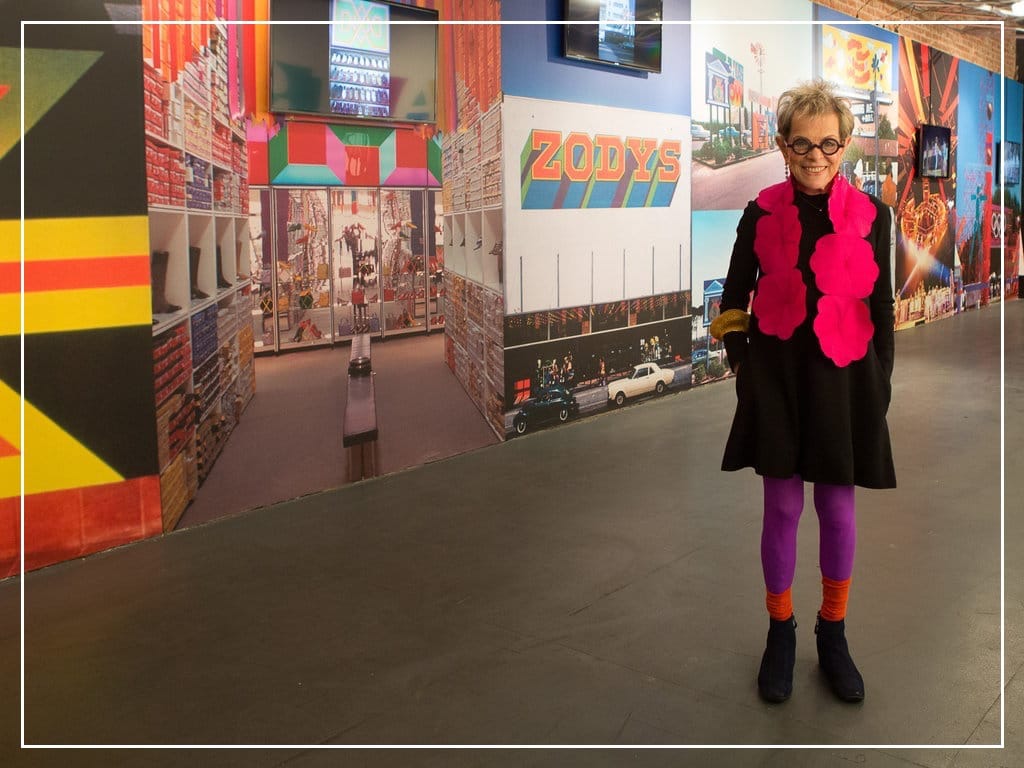 Designer Deborah Sussman, stylishly dressed in a hot pink felt boa, purple pants and orange socks, stands before a mural of her colorful graphic design work in a gallery