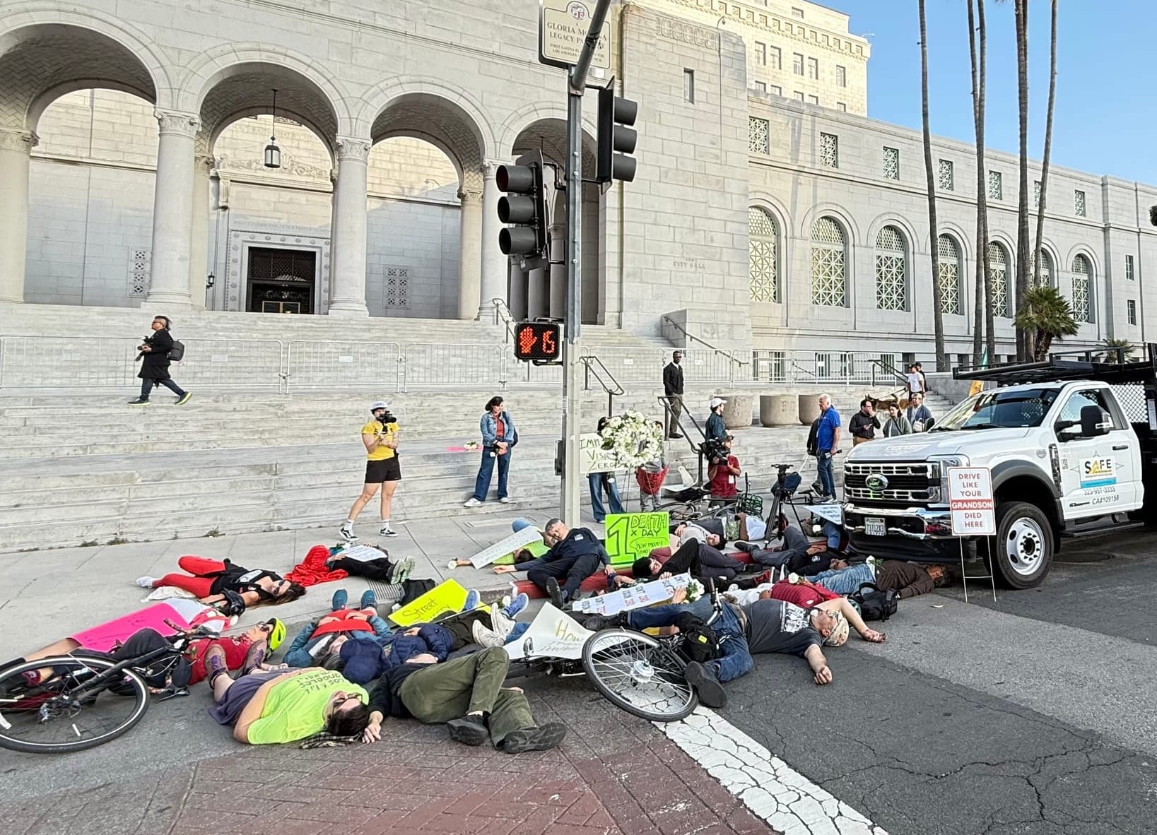 Another angle of the die-in showing dozens of bodies strewn on the ground in front of City Hall with a neon green sign that says one death a day