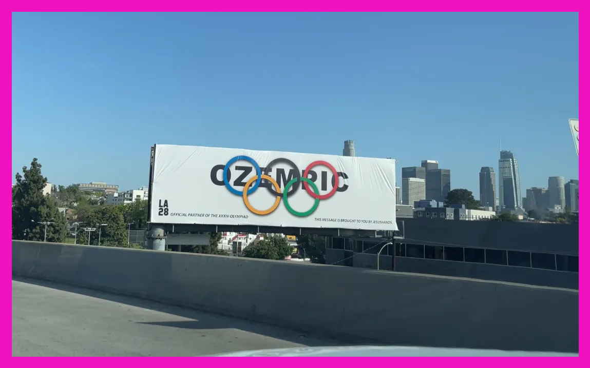 A billboard that says OZEMPIC with the Olympic rings installed in front of the downtown LA skyline