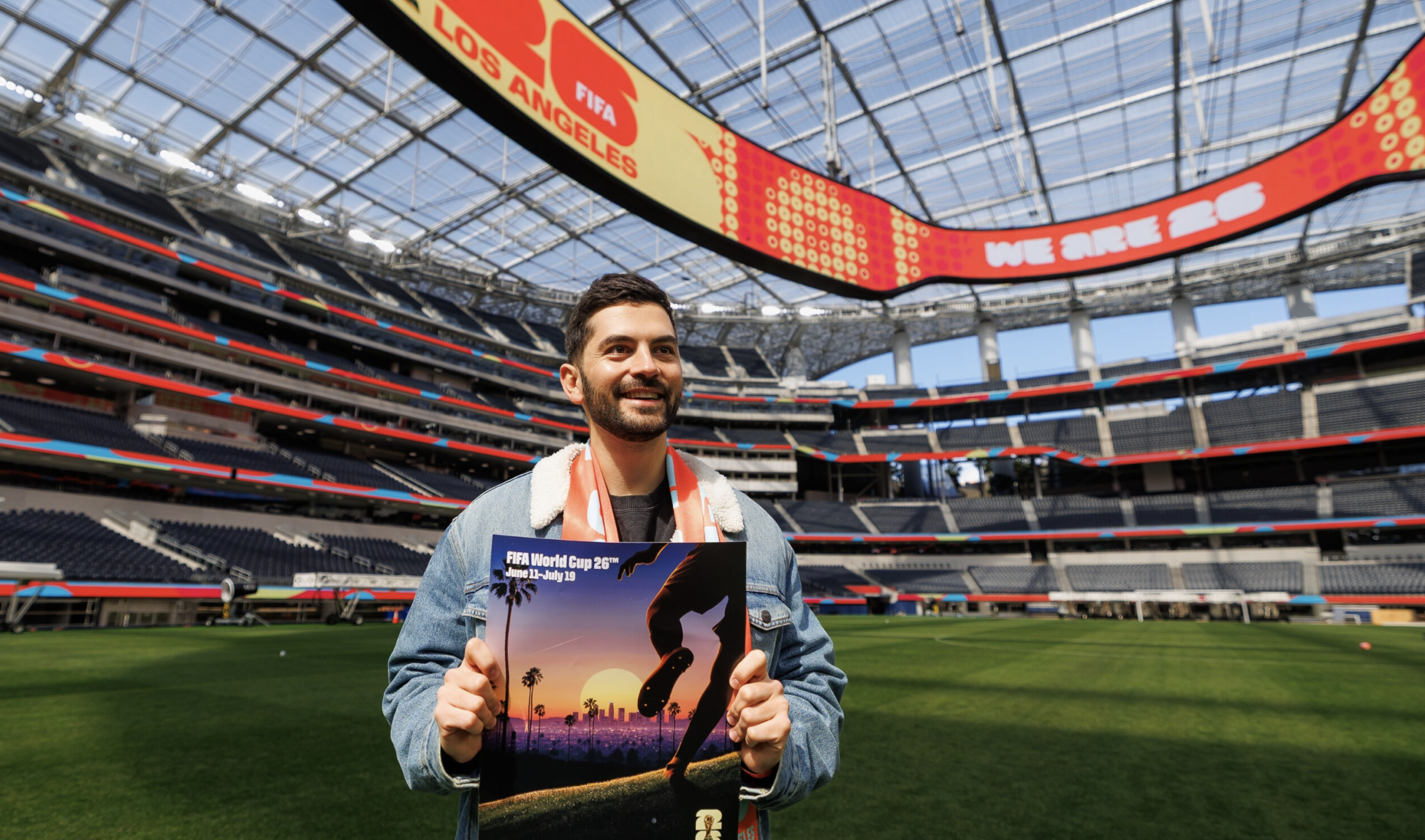 Artist Thieb Delaporte-Richard holds his poster for the LA World Cup tournament showing a soccer cleat against the sunset of downtown LA, while he stands in SoFi Stadium filled with LA World Cup graphics like WE ARE 26