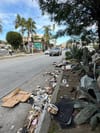 A street filled with trash in the gutter: wrappers, trash bags, cups, buckets, boxes, clothing, car parts
