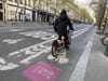 A person in a black coat rides a bike share bike in a pink Paris 2024-branded bike lane on a Paris street
